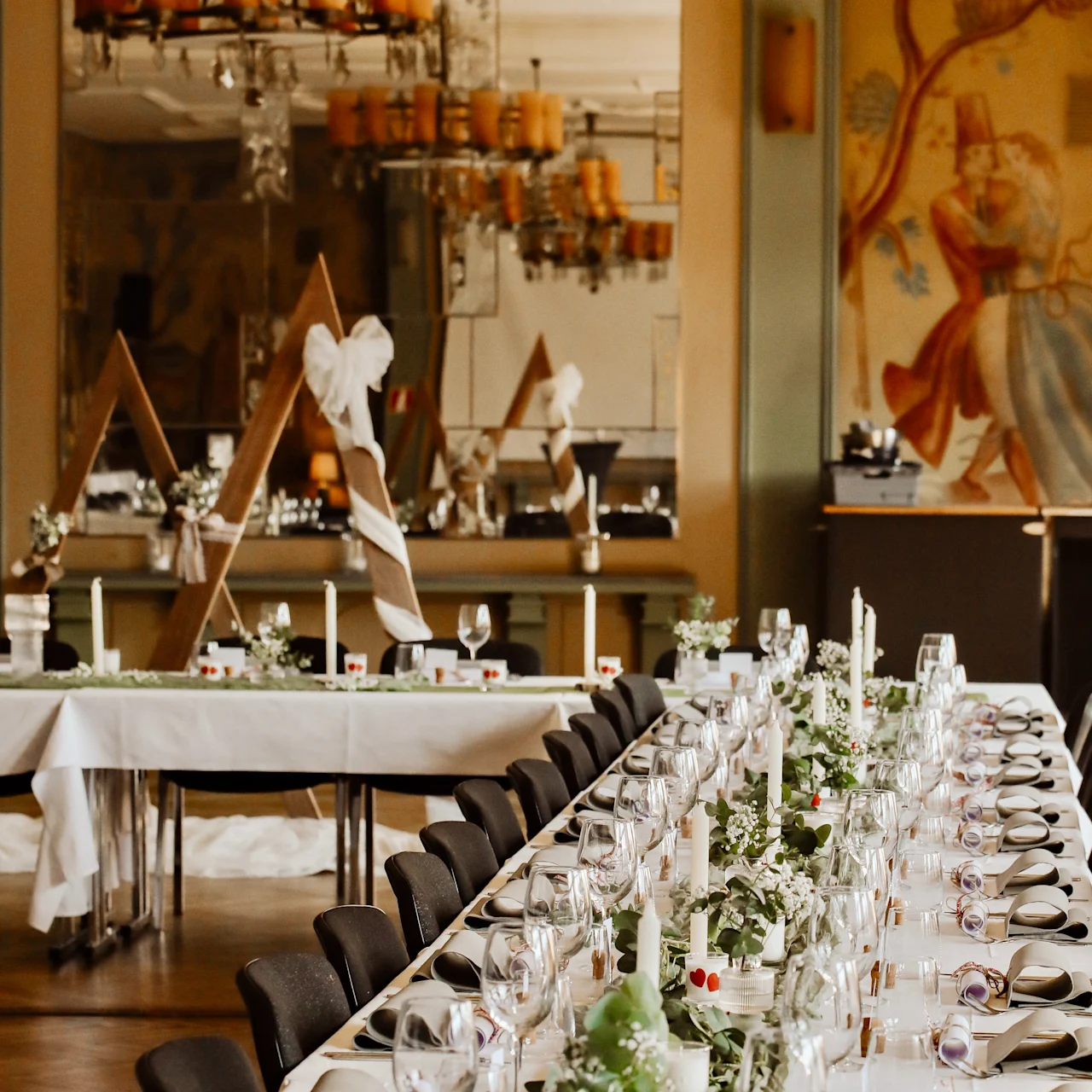 A long, elegantly set dining table with white tablecloths is decorated with green foliage, candles, and glassware. A mural and large chandeliers create a refined dining atmosphere.