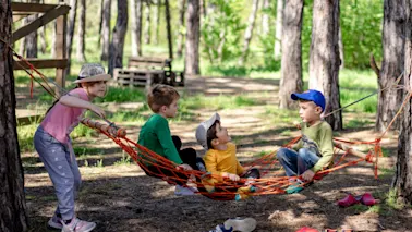 Barn leker i en hengekøye i en solrik skog. Ett barn dytter hengekøyen mens andre sitter og prater.