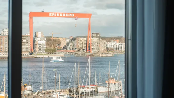 Näkymä ikkunasta satamaan, jossa on veneitä, kaupungin siluetti ja suuri punainen nosturi, jossa lukee ERIKSBERG.