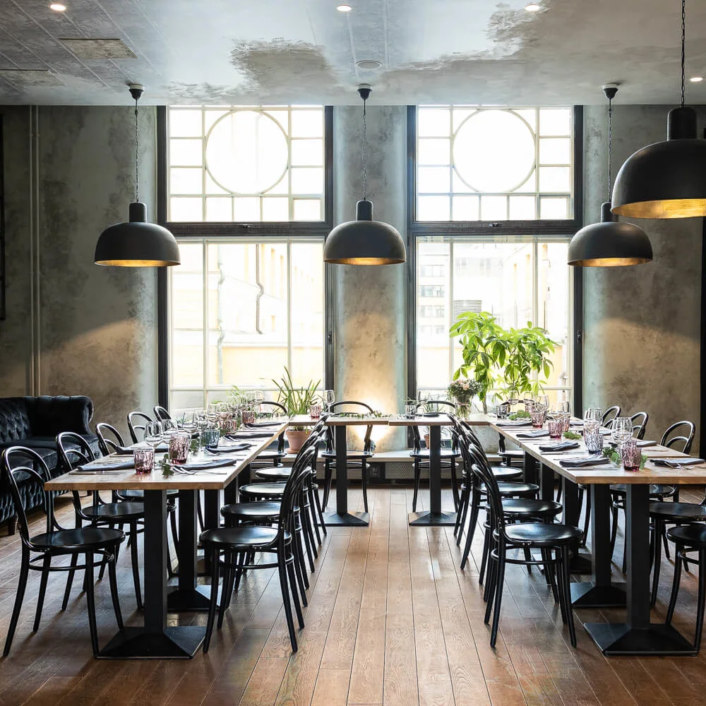Bord arrangert med servise og stoler, opplyst av hengende lys, i en stilfull restaurant med store vinduer og tregulv.