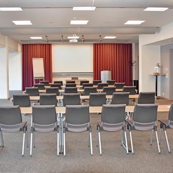 Rader av tomma stolar och skrivbord vända mot en vit skärm i ett väl upplyst konferensrum med röda gardiner, blädderblock och väggmonterad TV. En klädhängare och ett litet bord syns.