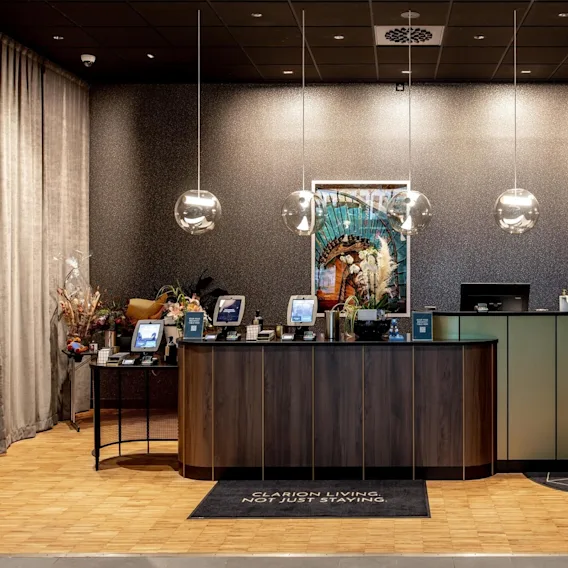 Reception desk features computers and hanging lights, surrounded by elegant decor and curtains. A carpet reads, "CLARION LIVING, NOT JUST STAYING." Artistic wallpaper adds a sophisticated touch.