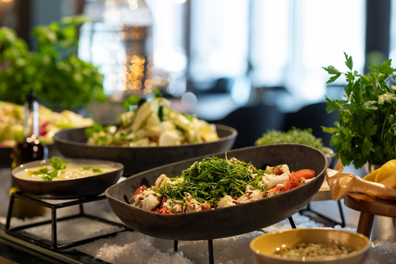 Skålar med färska sallader ställs ut på is, garnerade med örter. En suddig restaurangmiljö med växter och ytterligare rätter i bakgrunden.