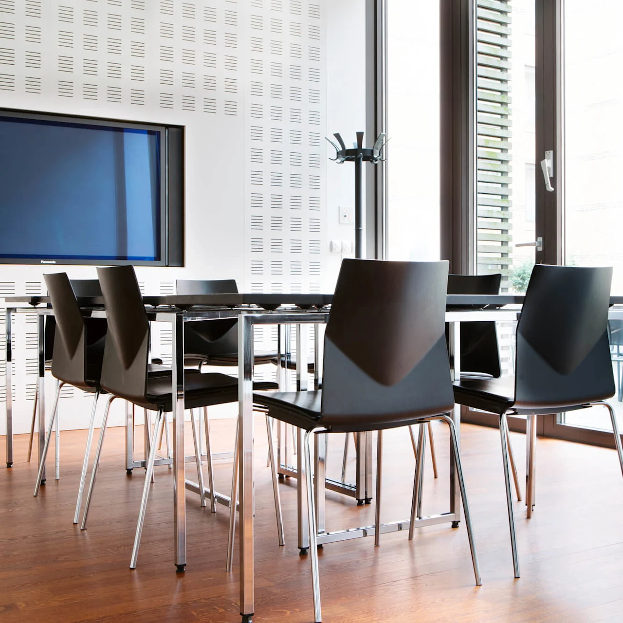 A modern meeting room with a large TV screen mounted on a perforated wall. A black meeting table with matching chairs is set on a wooden floor, surrounded by large windows allowing natural light to flood the room.