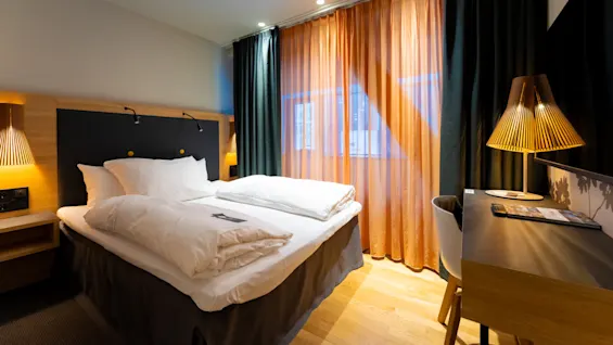 En seng med hvidt sengetøj står i et moderne hotelværelse. Blødt lys oplyser træpanelsvægge. Et skrivebord med lampe og stol er i nærheden.