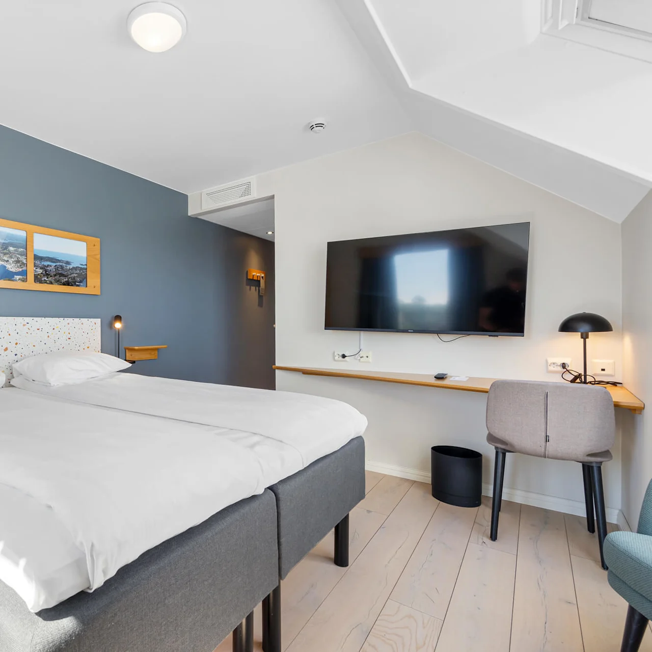A neatly made double bed in a modern hotel room, flanked by artwork, a desk, and a flat-screen TV.