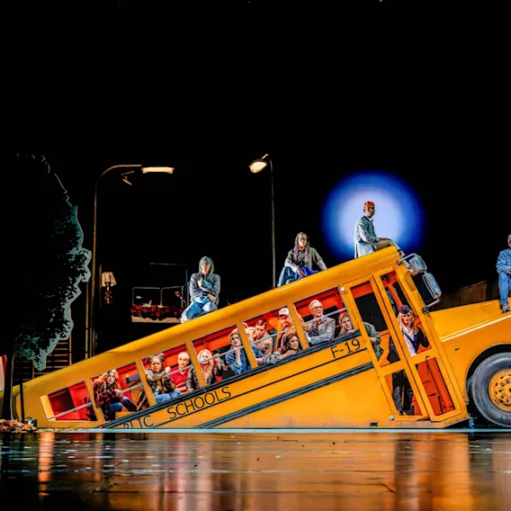 Teaterföreställning med skådespelare på och inuti en snedställd gul skolbuss