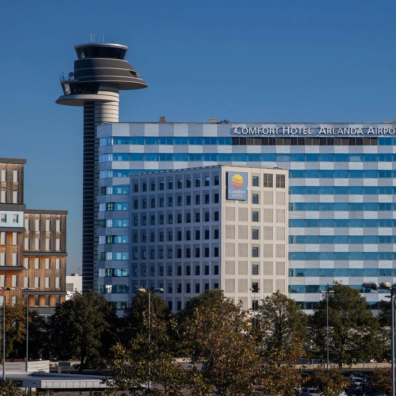 A large, modern hotel building labeled "Comfort Hotel Arlanda Airport" with reflective windows stands beside an airport control tower under a clear blue sky. Trees and other buildings surround the foreground.