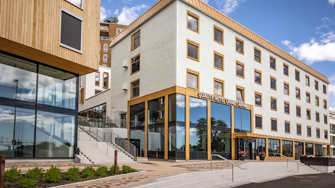 A modern hotel building with large windows and a minimalist design stands under a blue sky. The sign reads "QUALITY HOTEL GRAND LARVIK." Stairs and plants are in front.
