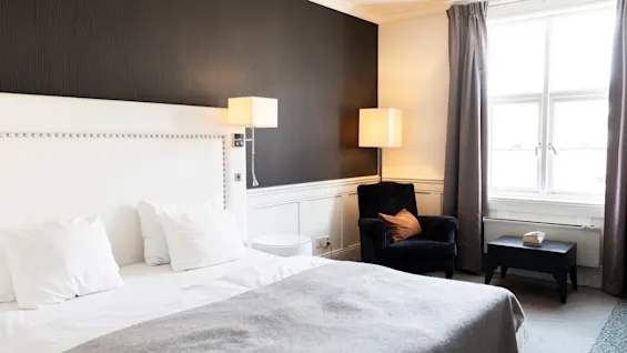 Et indbydende hotelværelse med en plys seng, en komfortabel lænestol og blød belysning, der skaber en rolig atmosfære.