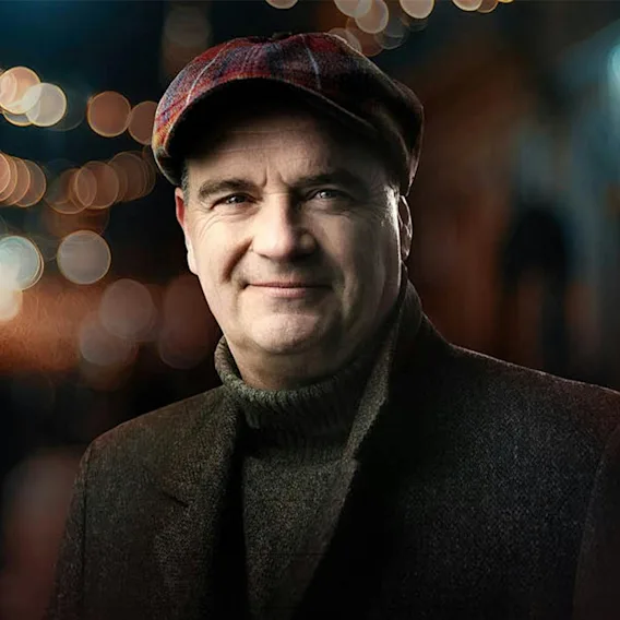 En smilende mann i tidsriktig lue og frakk som ser fremover mot en stemningsfull, glitrende kveldsbakgrunn.