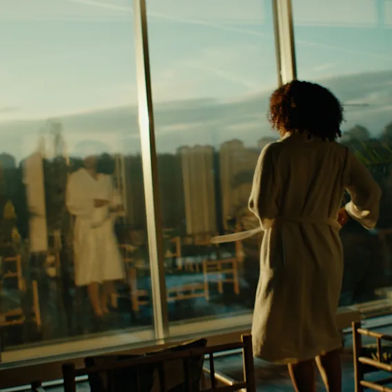 A person in a white robe stands indoors, gazing out through large windows at a cityscape during sunset, with plants nearby.