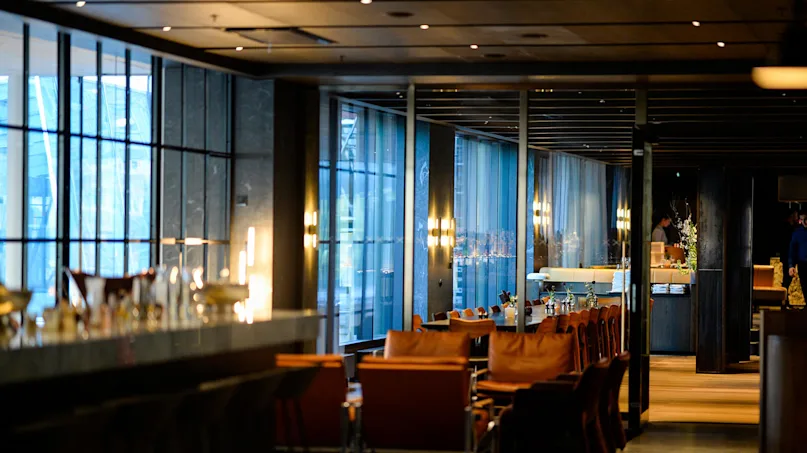 Chairs and tables are arranged in a dimly lit, modern restaurant with large windows and sleek decor, creating a sophisticated dining atmosphere.