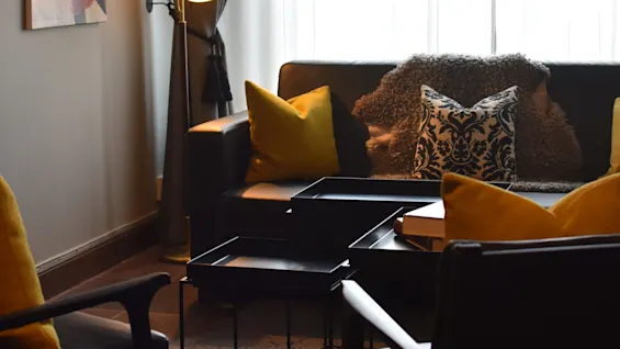 En hyggelig stue har en moderne sofa med gule og mønstrede puder, oplyst af en gulvlampe. Blødt lys filtreres gennem tynde gardiner, med et farverigt maleri på væggen.