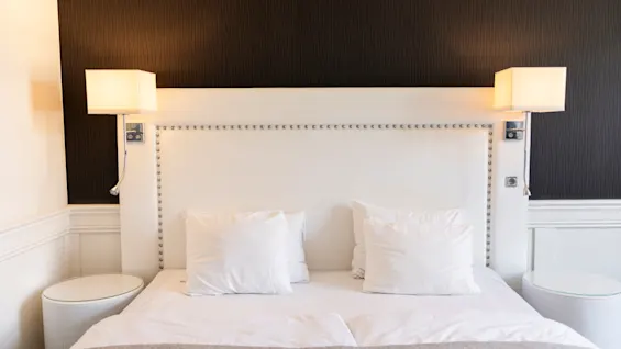 Et hyggeligt hotelværelse med en pænt redt seng, hvid sengegavl og blød belysning fra vægmonterede lamper, der indbyder til afslapning.