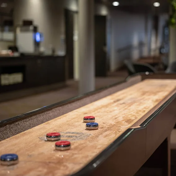 Et shuffleboard-bord med røde og blå pucker står i et loungeområde, omgitt av stoler og bord, som skaper en avslappet spillatmosfære.