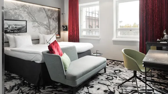 A modern hotel room with two beds, a sofa, and elegant decor illuminated by daylight from windows.