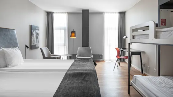 A hotel room with a neatly made double bed and a bunk bed; features chairs near tall windows, a desk with a red chair, and wood flooring.