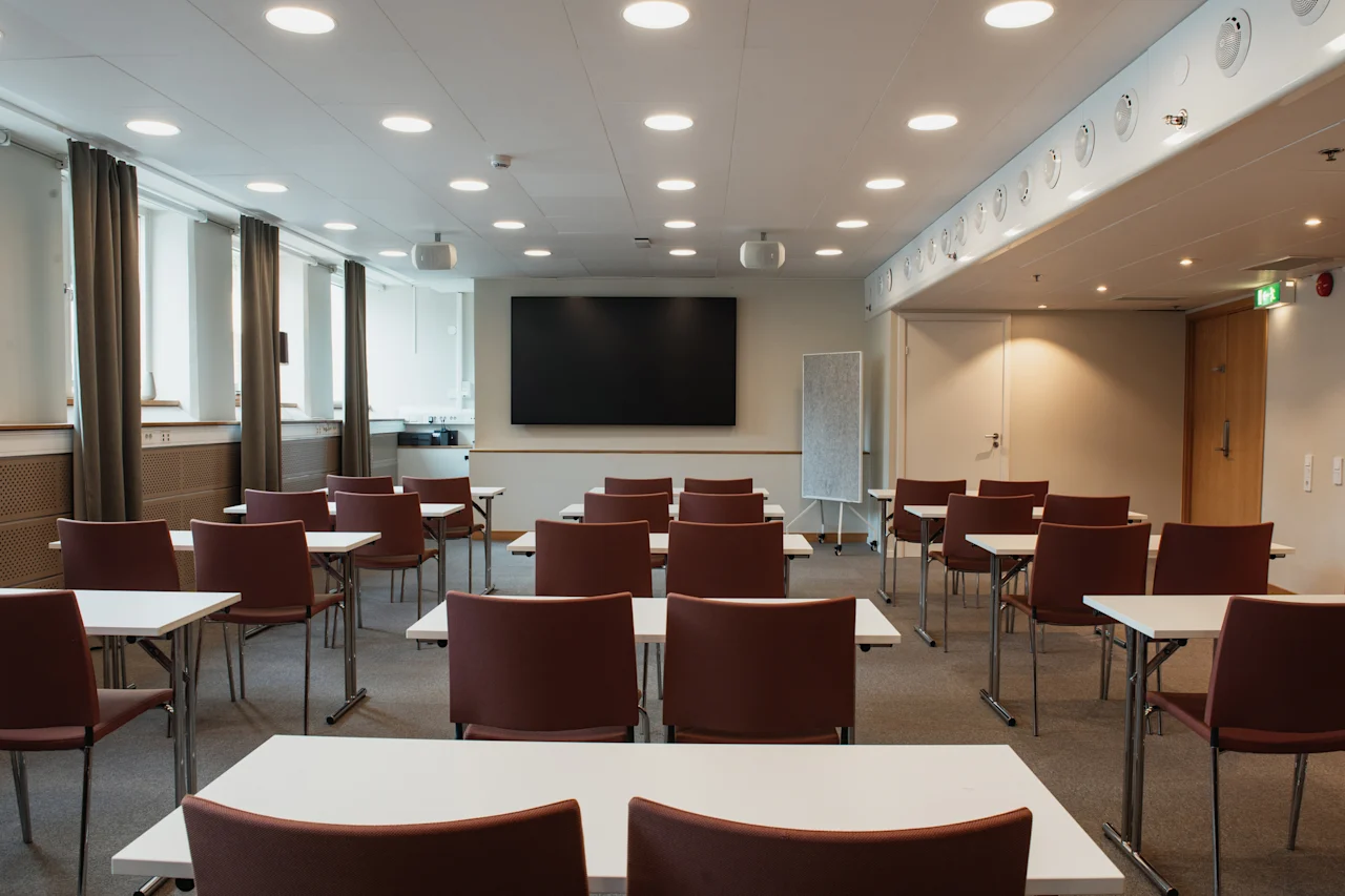 A well-lit conference room at Home Hotel Uppsala with tables and chairs set up for a presentation, facing a large screen.