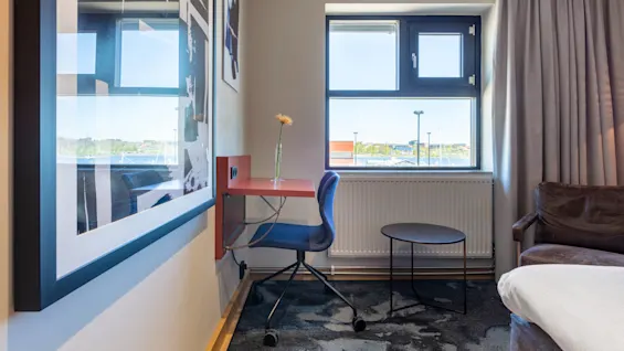 A well-lit room with a work desk and chair, a bed, window with a view, wall art, and curtains.