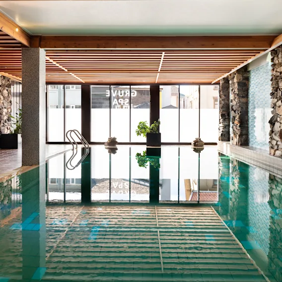 Innbydende innendørsbasseng på Quality Hotel Grand, Kongsberg, med naturlig lys, steinvegger, koselig sittegruppe og Grand Spa-refleksjoner.