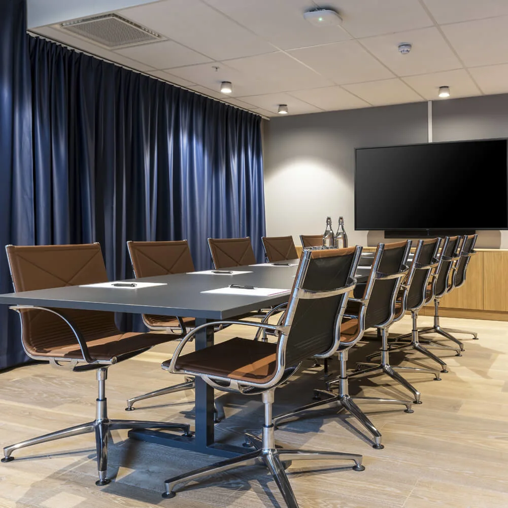 A long conference table surrounded by chairs sits in a room with blue curtains and a mounted TV. The floor is wooden, and the ceiling has spotlights.