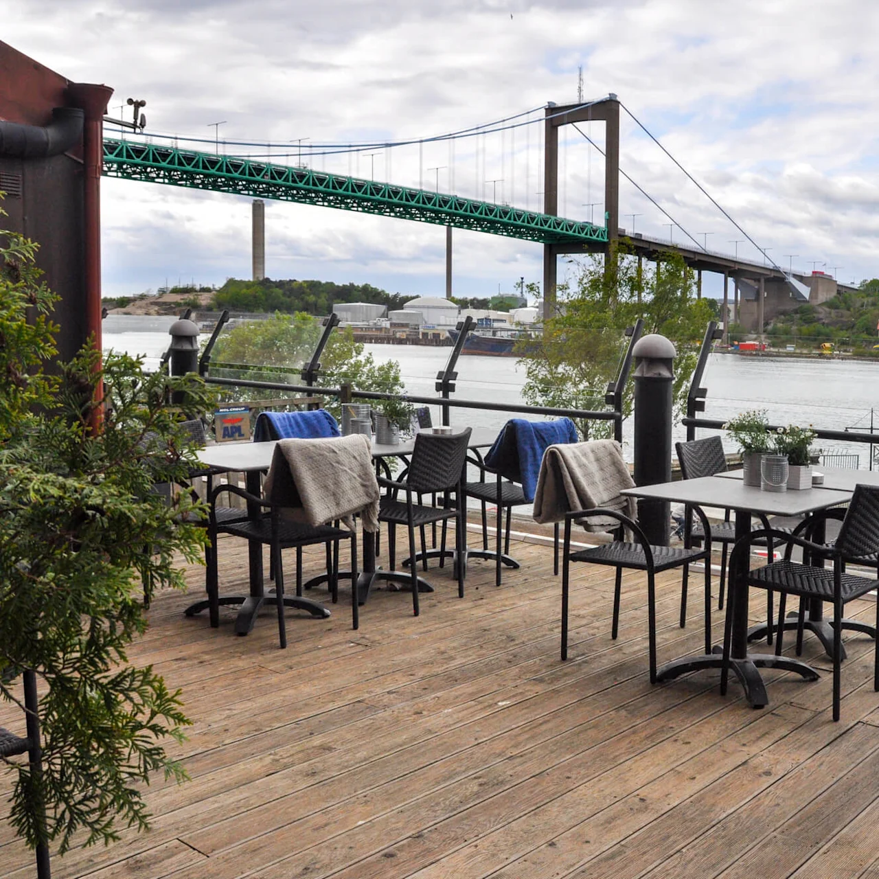 Outdoor café with empty tables and chairs offering blankets against a scenic river backdrop; a large green suspension bridge spans the water under a partly cloudy sky.