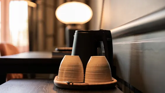 En sort elkedel står på et træbord ved siden af to beige kopper på en bakke. En blødt lysende lampe i baggrunden skaber en varm, indbydende atmosfære.