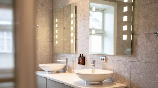 To moderne hvite vasker med kromkraner står på en beige benkeplate under opplyste speil. Grå flislagte vegger og et vindu gir et lyst, elegant baderomsmiljø.