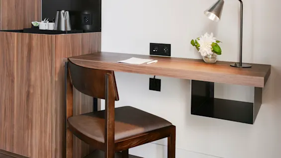 Ett skrivbord i trä med en metallampa och en blomvas står mot en vit vägg. En stol står under, och hyllor med glas och en vattenkokare finns i närheten.
