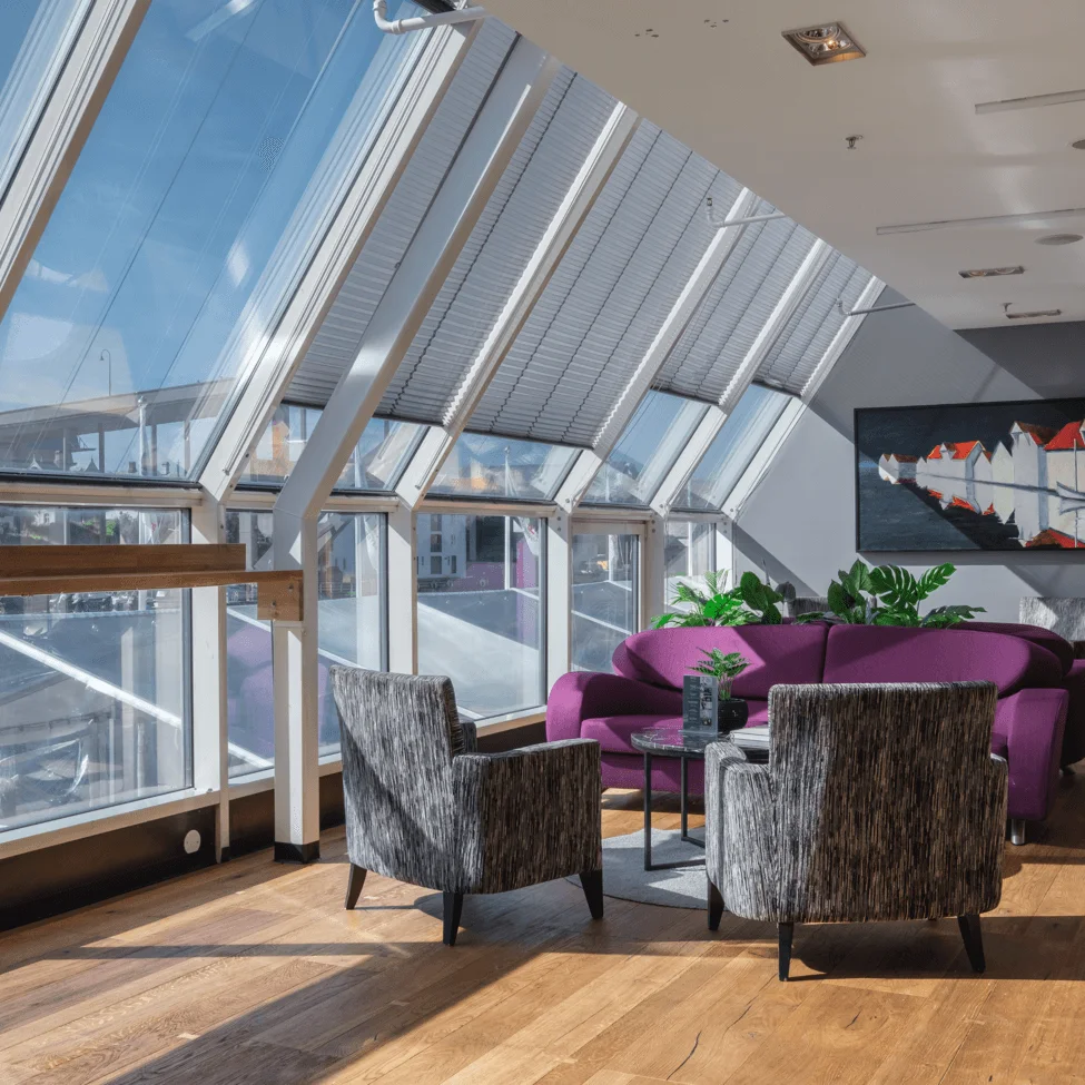 Lila soffor och grå fåtöljer omger ett lågt bord i ett solbelyst rum med lutande glasfönster. En krukväxt och en modern målning förbättrar den mysiga atmosfären.