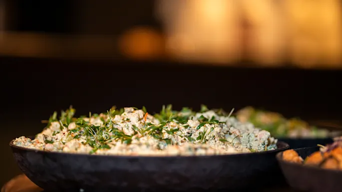 A black bowl holds herbed rice garnished with fresh greens, set on a wooden table in a softly lit dining area.