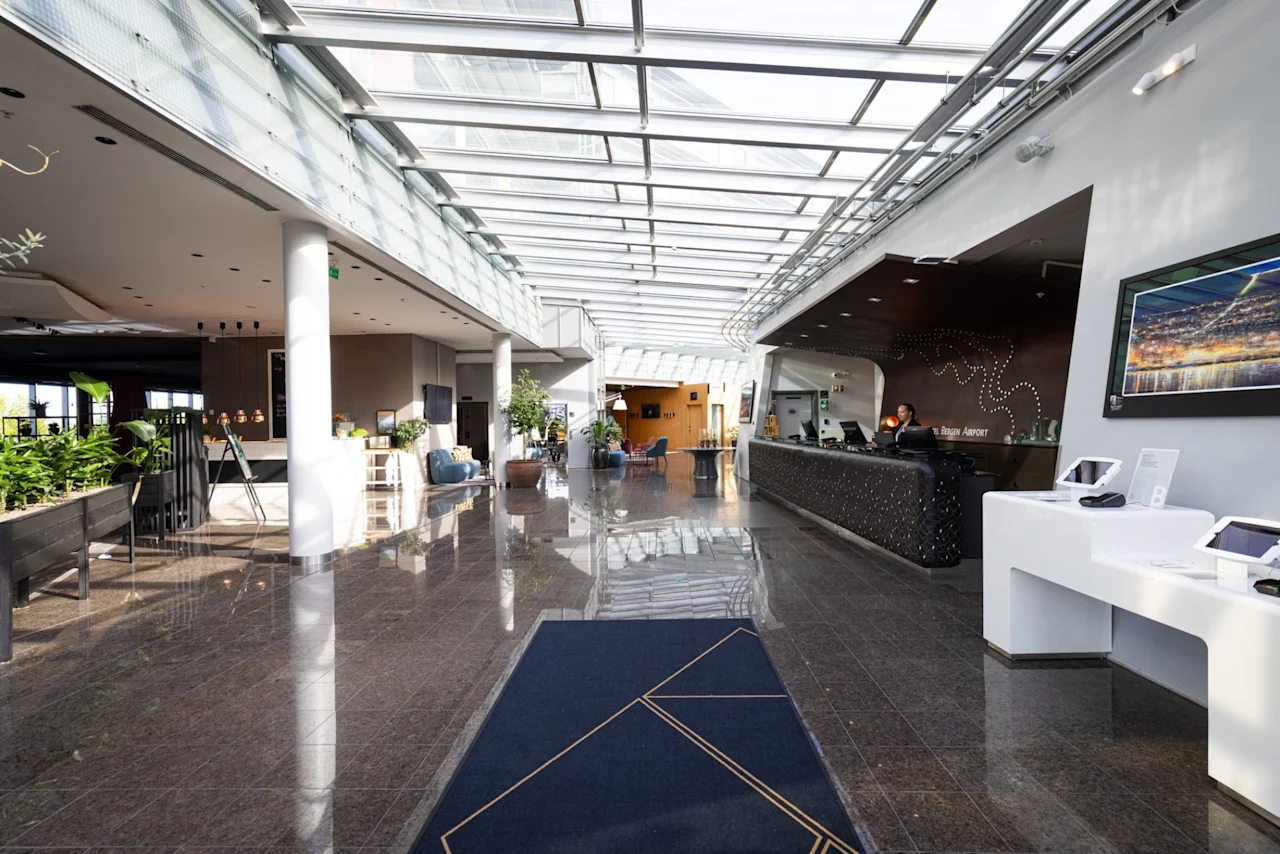 En hotellobby med glinsende fliser. En resepsjonist står bak en svart skranke til høyre og ønsker gjester velkommen. Potteplanter og moderne møbler pryder den romslige, solfylte korridoren.