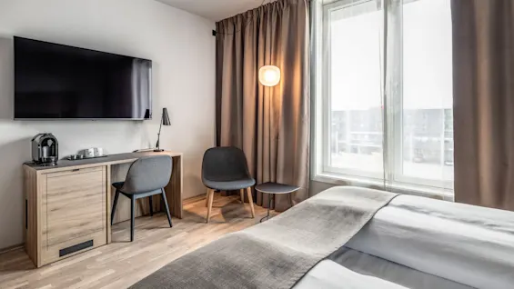 Et moderne hotelværelse med en seng, en vægmonteret tv og et skrivebord med kaffemaskine. Et stort vindue med gardiner og en lampe giver naturligt lys.