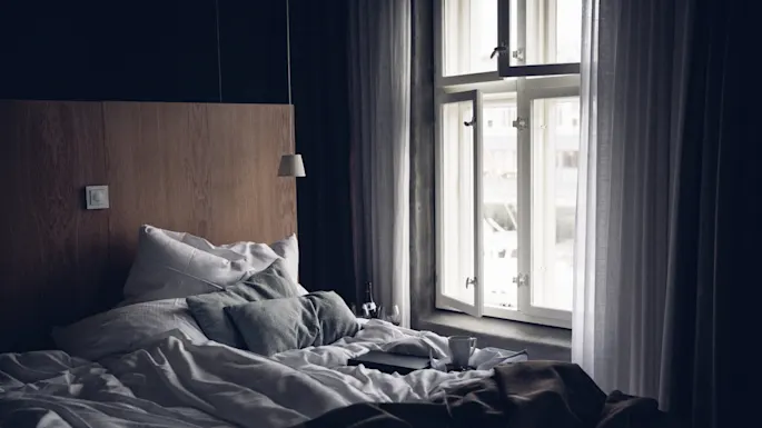 A neatly made bed with white and gray pillows, brown blanket, and open windows; a mug and closed book lie atop. The room appears calm and softly lit.