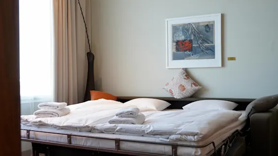 A neatly made bed with folded towels on top, positioned in a hotel room beside a window with closed curtains; a framed artwork and decorative pillows adorn the wall.