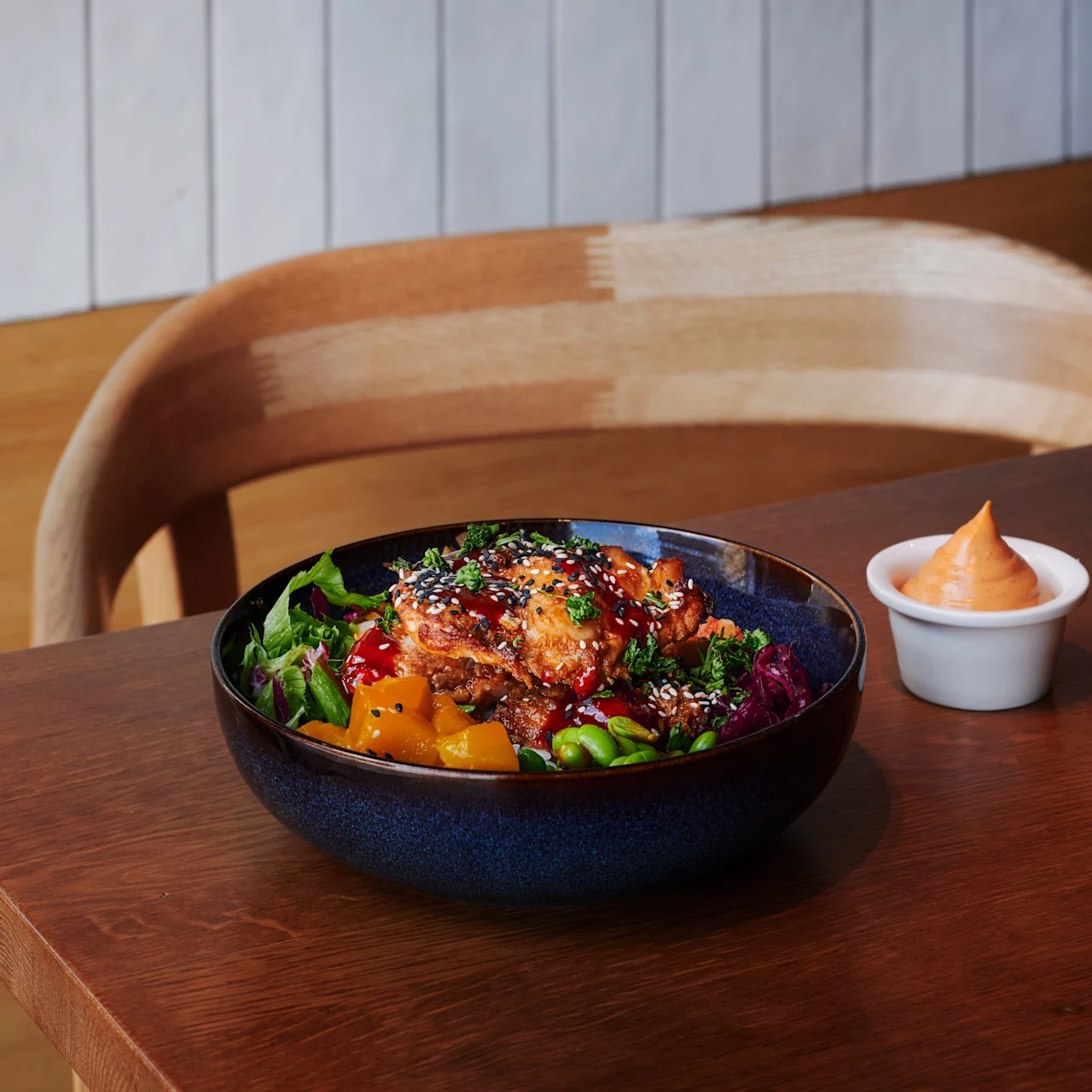A bowl filled with colorful salad and topped with sesame seeds sits on a wooden table beside a small dish of orange sauce, against a light wooden wall and chair backdrop.