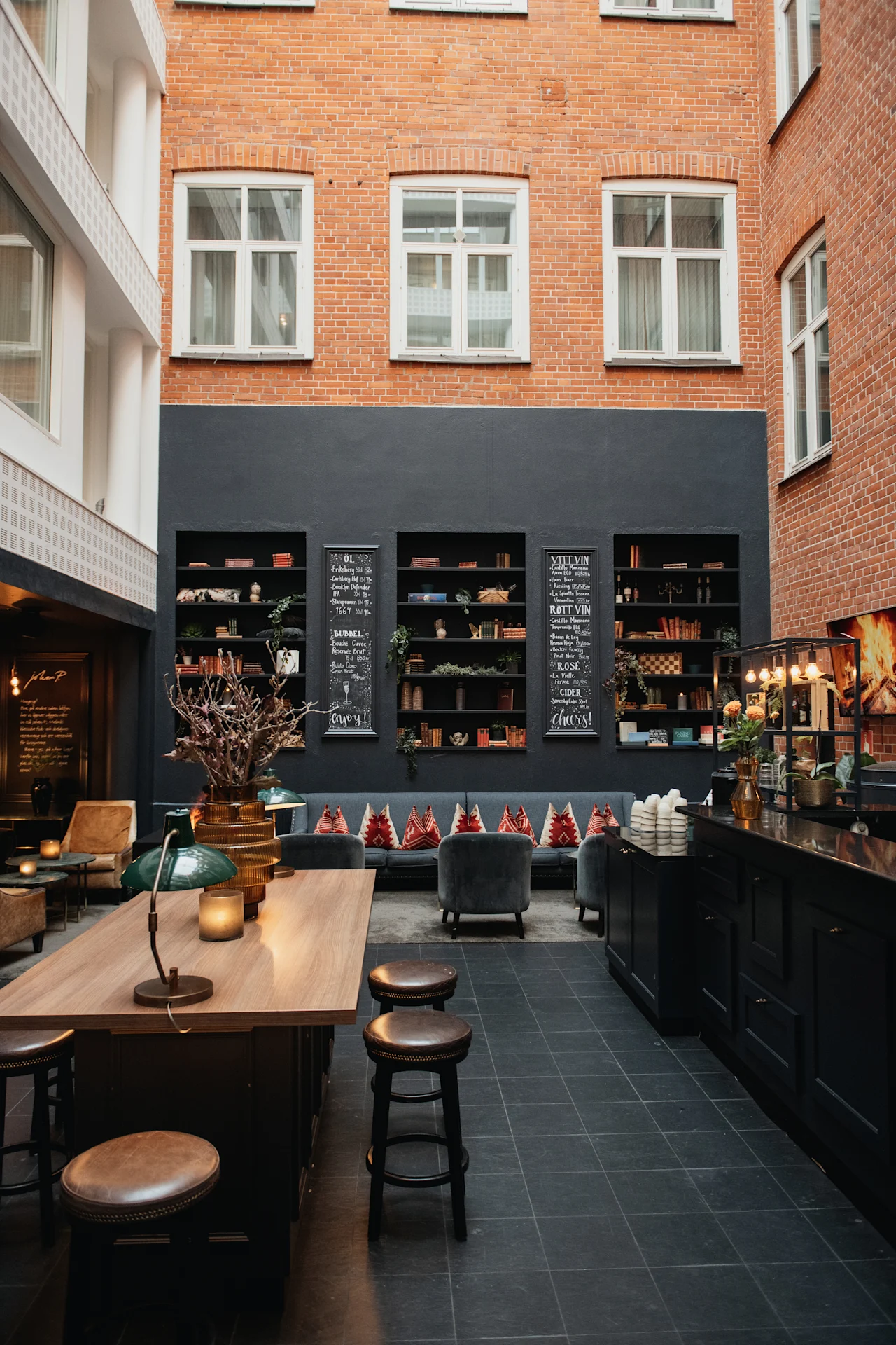 En hyggelig hotellounge med murstensvægge, bogreoler, et langt træbord og en bar. Menuen inkluderer BUBBEL, VITT VIN, ROTT VIN, ROSE, CIDER. Nyd og skål!