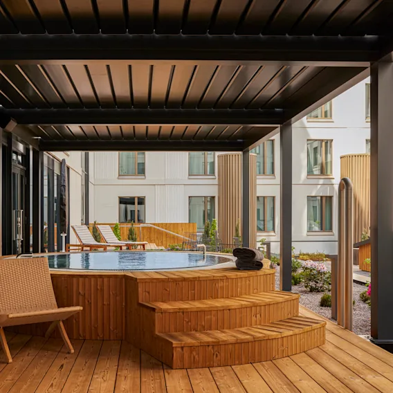 Puinen poreallas sijaitsee terassilla modernin pergolan alla. Lähellä on tuoleja ja pöytiä luomassa rentouttavaa terassiympäristöä, jota ympäröivät suuret ikkunat omaavat modernit kerrostalot.