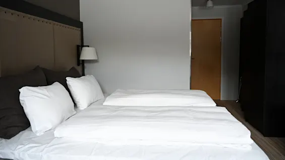 En snyggt bäddad säng med vita lakan och kuddar står mot en taupe-sänggavel. En lampa är monterad på väggen. En träfärgad dörr och garderob syns.