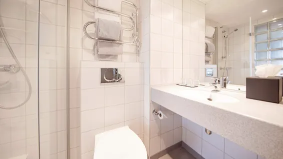 A white-tiled bathroom features a wall-mounted toilet and sink. Towels hang on a heated rack beside a shower with glass doors, creating a clean, modern atmosphere.