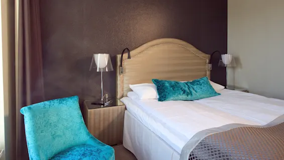 An elegant bed with a beige headboard flanked by modern lamps, accented by vibrant blue pillows and chair in a stylish hotel room.
