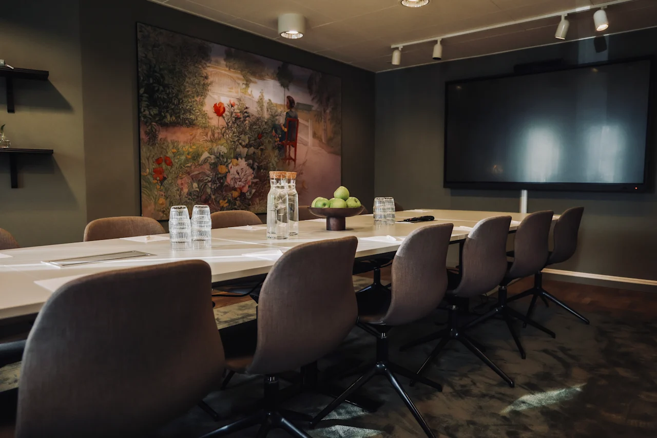 A long conference table with chairs holds pitchers, glasses, and a bowl of green apples. In the room are a large screen and a colorful painting.  