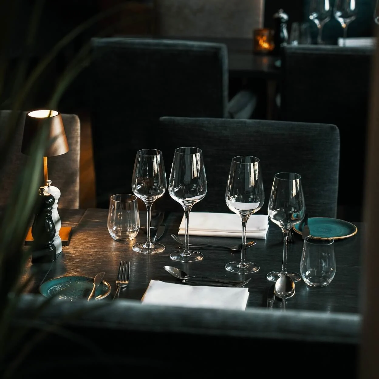 Et spisebord, arrangert med vinglass og bestikk, står i en svakt opplyst restaurangmiljö. En liten lampe gir myk belysning.