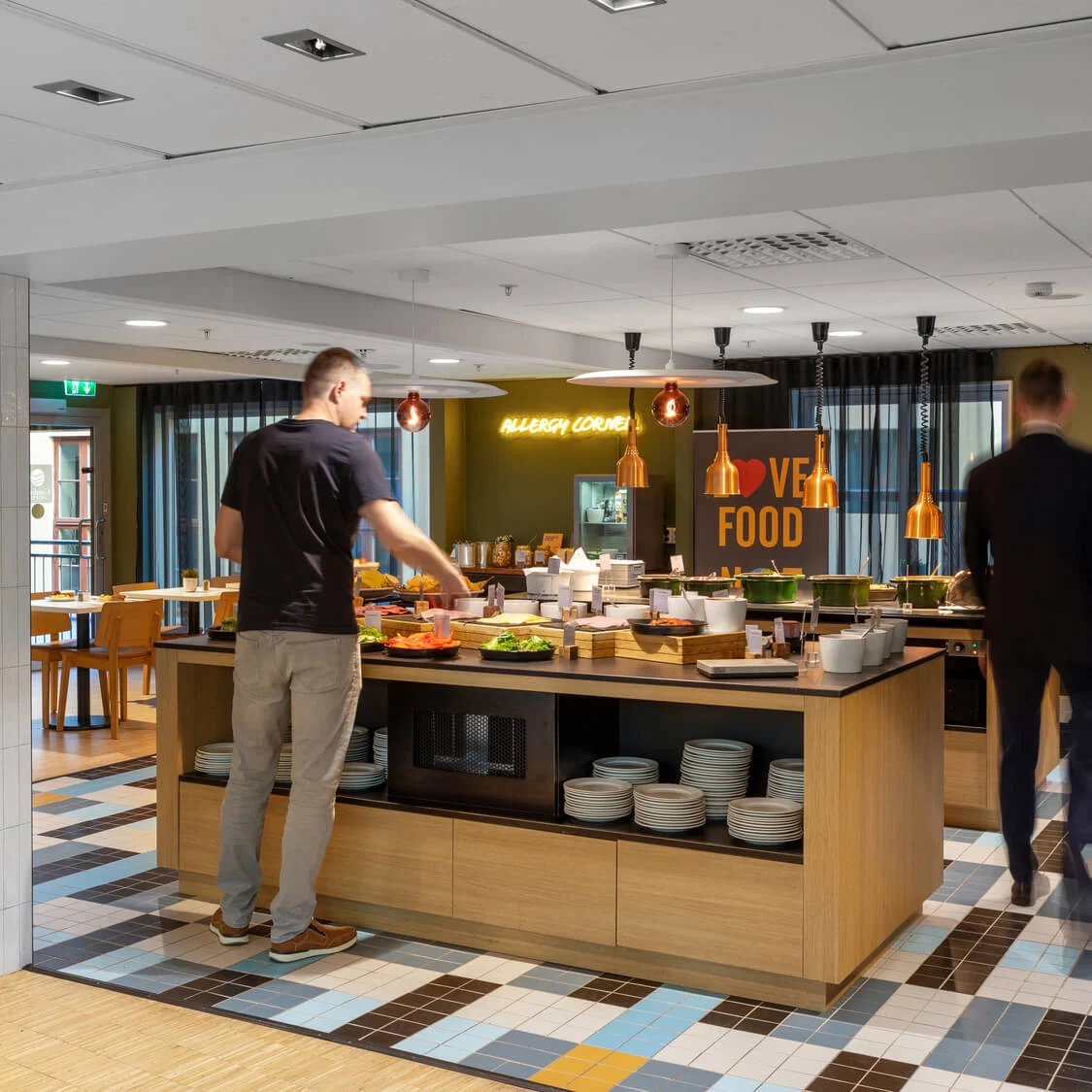 En man tar för sig vid en buffédisk i en modern kafeteria med träinredning och färgglada kaklade golv. Skyltar säger "ALLERGY CORNER" och "LOVE FOOD".