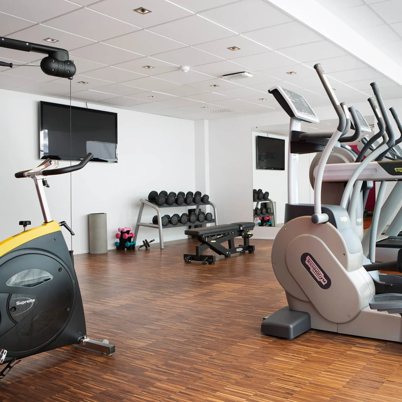Treadmills and exercise bike sit unused in a gym, surrounded by dumbbells and benches on a wooden floor with a wall-mounted TV.