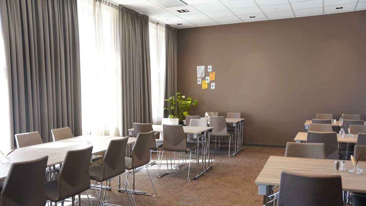 Tables and chairs arranged in a modern conference room with large windows, curtains, and a potted plant.