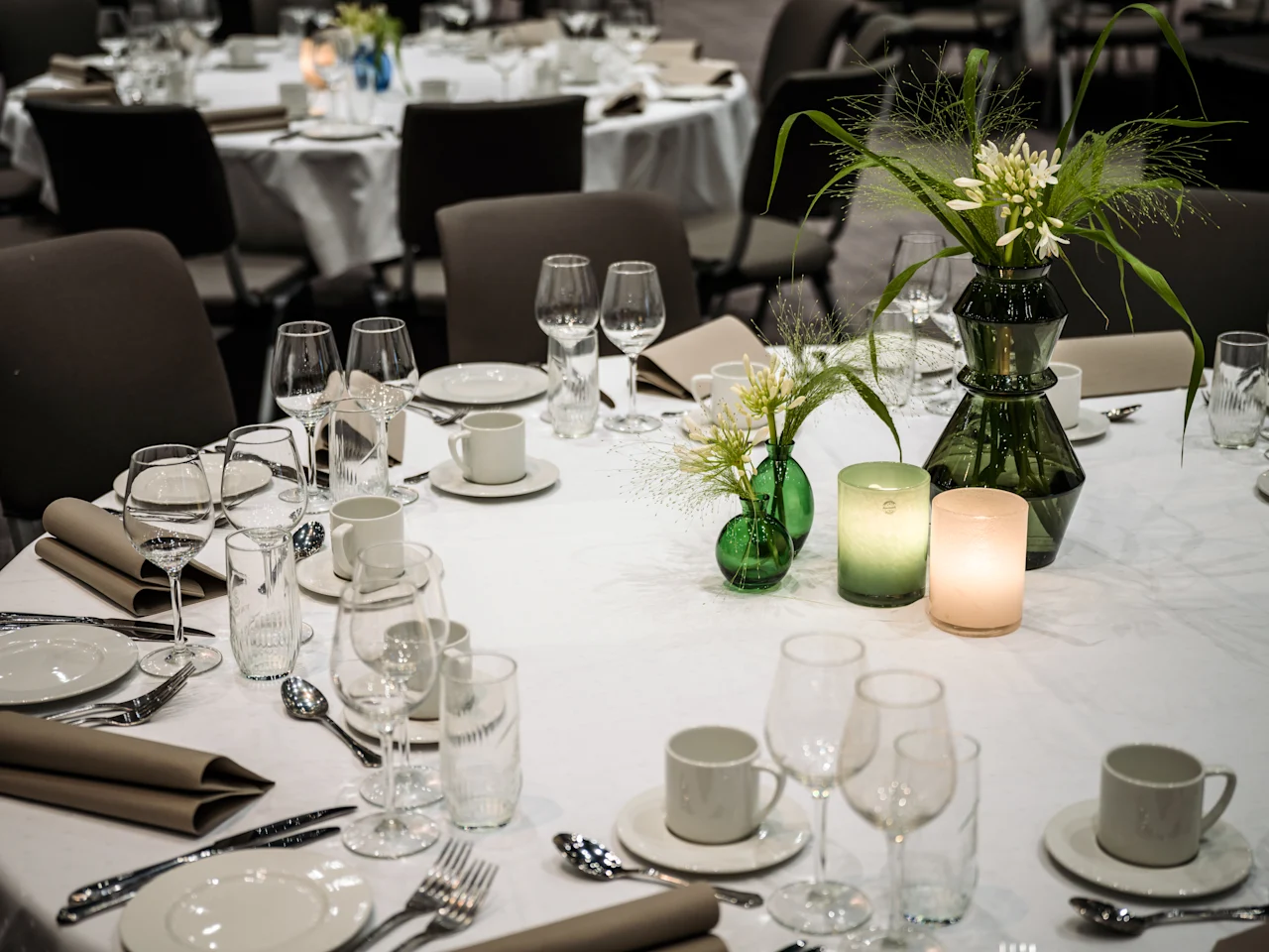 Ett formellt middagsbord dukat för ett evenemang, med flera kuvert, gröna blomsterarrangemang och glödande ljus som skapar en varm atmosfär.