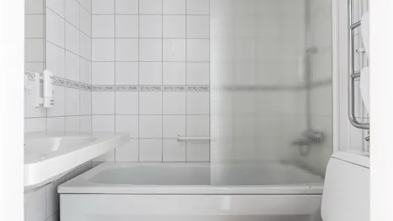 Badkar med frostat glas, mot vita kakelväggar. Intilliggande handfat och toalett kompletterar det kompakta badrummet. En liten handdukstork är monterad i närheten.