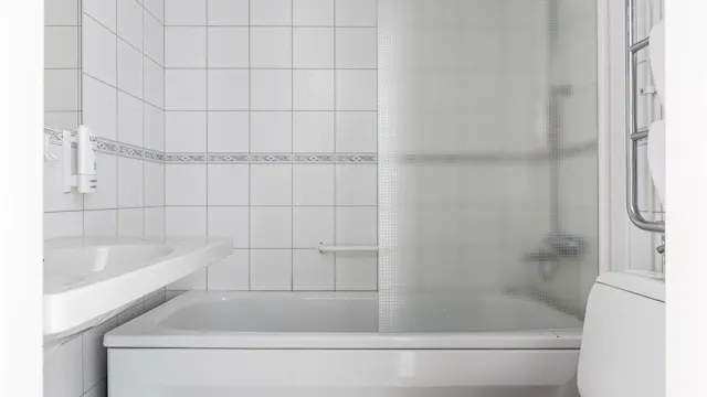 Badkar med frostat glas, mot vita kakelväggar. Intilliggande handfat och toalett kompletterar det kompakta badrummet. En liten handdukstork är monterad i närheten.